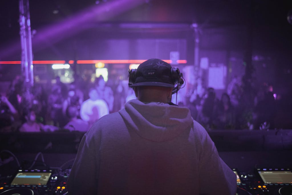 DJ in hoodie playing live set at a vibrant nightclub with purple lighting.